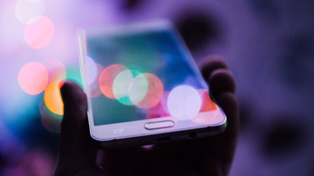 カラフルなボケ光の中でスマートフォンを持つ手。モバイル通信のイメージ